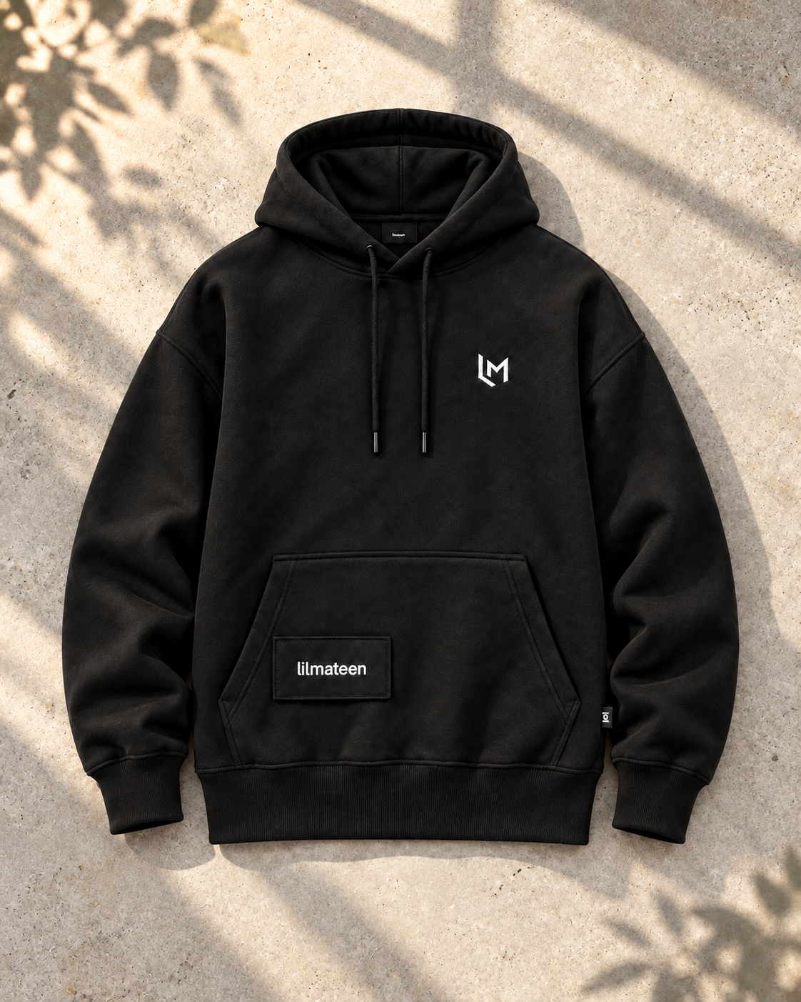 Black LILMATEEN UNFIXED 001 hoodie on sunlit concrete with soft leafy shadows